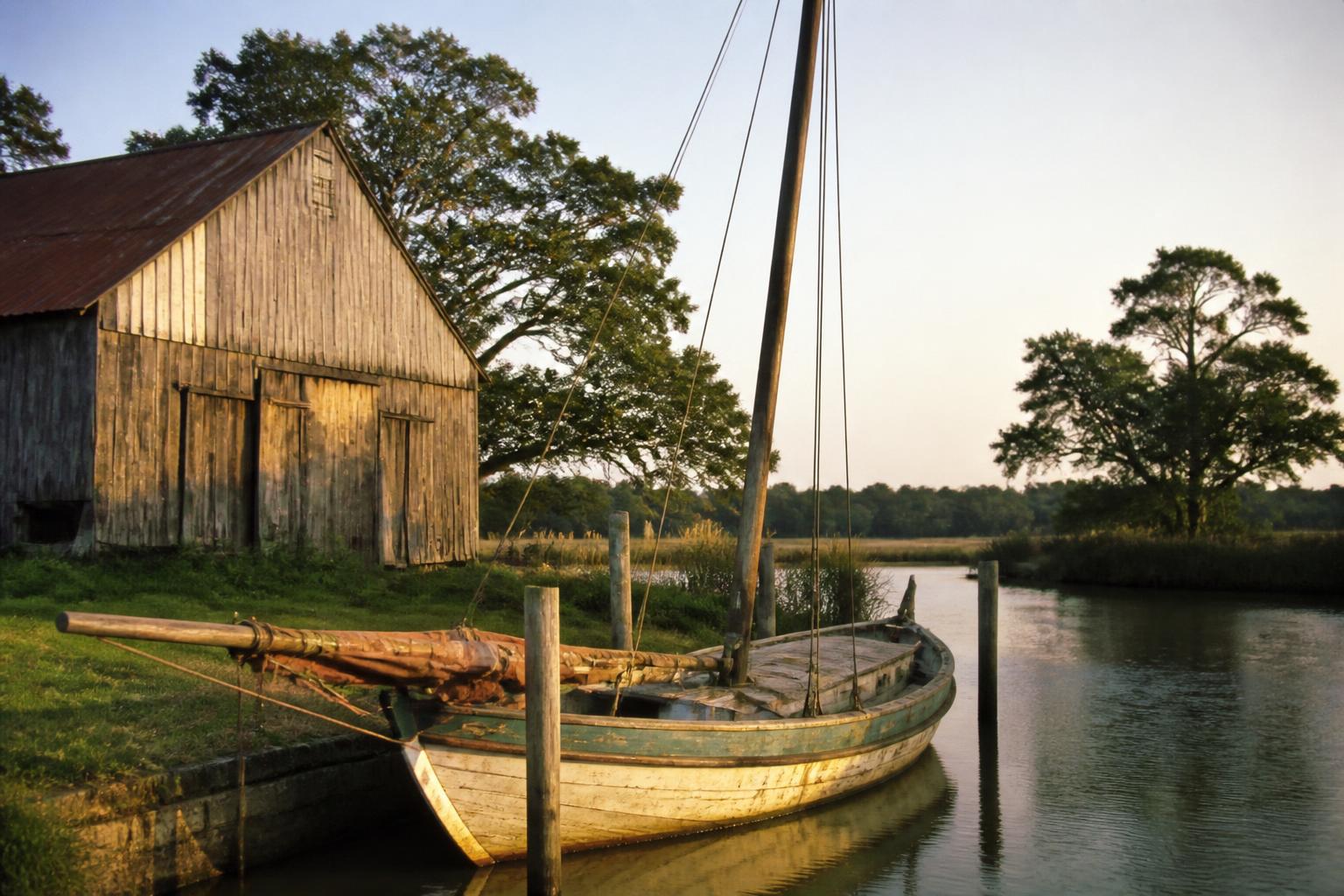 Weathered tobacco barn beside a wooden sailing workboat on a tidal creek